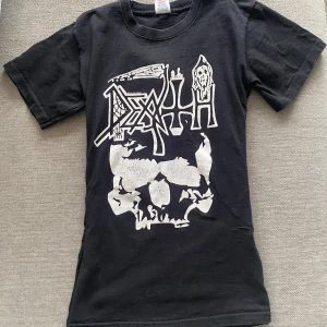 Svart t-shirt med dödskalletryck - Cool svart t-shirt med ett stort tryck av en dödskalle och texten 'Death' i vitt. Perfekt för dig som gillar rock eller metal. T-shirten har korta ärmar och en klassisk rund halsringning. Insydd för sitta tight men går enkelt att sprätta up (från 2009)