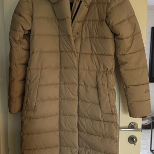 Beige lång dunjacka - Säljer en snygg beige lång dunjacka med huva. Jackan har en stilren design med dragkedja och knappar framtill. Perfekt för kyliga dagar med sin långa passform och quiltade mönster.! 🧥 Didrikson i storlek 34, den är för stora för mig, skulle säga att den nog passar upp till en liten 38/M beroende på vart man drar. Har knappt använt dem så den är som nya.  Yttermaterial 100% polyester.  Längd 121 cm rakt ner över ryggen Bröst bredd 50 cm  Tvättas 40 grader skon tvätt 