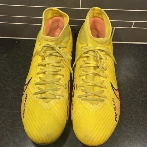 Gula Nike fotbollsskor - Säljer ett par gula Nike fotbollsskor med snörning och hög ankel. Skorna har en iögonfallande design med svarta och orange detaljer. Perfekta för fotbollsplanen med sina dobbar för bra grepp.