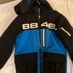 Svart och blå jacka från 8848 Altitude - Säljer en cool svart och blå jacka från 8848 Altitude med stora vita siffror på bröstet. Jackan har en dragkedja framtill och flera praktiska fickor. Perfekt för kyliga dagar med sin quiltade insida och justerbara huva.