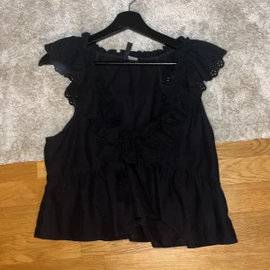 Svart ärmlös blus med spetsdetaljer - Snygg svart ärmlös blus med vackra spetsdetaljer runt halsen och axlarna. Perfekt för en stilren look.