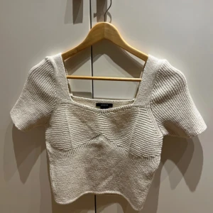 Vit stickad topp från Lindex - Säljer en stilren vit stickad topp från Lindex. Toppen har korta ärmar och en fyrkantig halsringning. Den är ribbad och har en figurnära passform som ger en snygg siluett. Perfekt för att klä upp eller ner beroende på tillfälle.