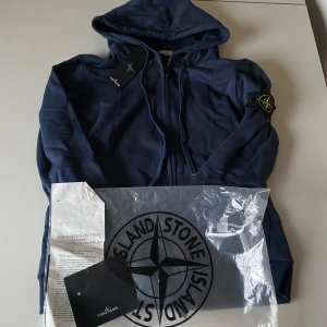 Blå hoodie från Stone Island - Säljer en snygg blå hoodie från Stone Island med dragkedja och huva. Tröjan har en ikonisk logga på ärmen och ribbade muddar vid ärmslut och nederkant. Perfekt för en stilren look.