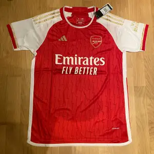 Säljer en ny Arsenal fotbollströja i rött och vitt från Adidas. Tröjan har korta ärmar och ett snyggt mönster med klubbens emblem och sponsorlogga på framsidan. Storlek M & perfekt för alla Arsenal-fans! 🏆