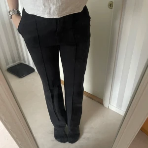 Svarta kostymbyxor - Säljer ett par stilrena svarta kostymbyxor. De har en klassisk design med knappar och dragkedja framtill. Perfekta för både formella tillfällen och vardagsbruk.