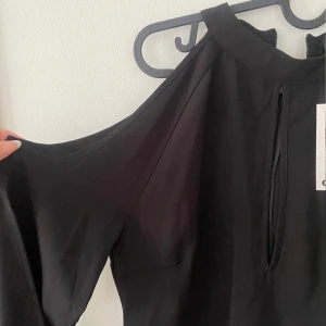 Boohoo svart blus, storlek M - Snygg svart blus med öppna axlar. Blusen har en hög hals och långa ärmar. Den är perfekt för en festlig tillställning eller en kväll ute.