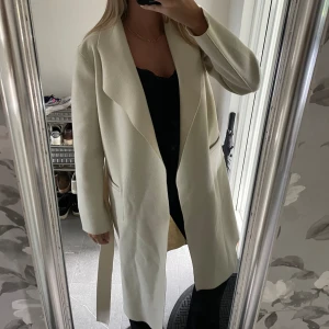 Lång kappa  - Säljer en super fin lång créme vit/ beige kappa från Vila. Kappan kan vändas ut och in om man vill få ett annat stuk och modell med sömmar! Det finns två fickor i fram samt ett band i midjan. Perfekt jacka till hösten😍😍
