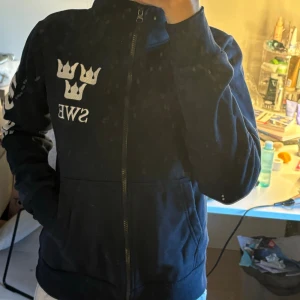 Svart jacka med dragkedja och tryck - Säljer en marinblå tjock ridtröja med dragkedja och coolt tryck på bröstet och ärmen. Jackan har långa ärmar och en hög krage. Perfekt för en avslappnad stil!