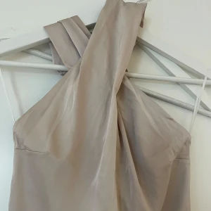 Beige satinklänning - Satinklänning i beige med halterneck och omlott vid benen. Knyts bak i nacken 