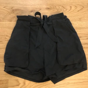 Knytshorts från Monki - Svarta tygshorts med knytning i midjan. Nästan oanvända och därmed i gott skick. Storlek 38. Normal till hög midja.