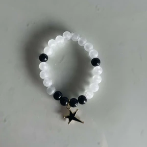Armband med stjärnhänge - Snyggt armband med vita och svarta pärlor. Det har ett coolt stjärnhänge i guld som ger en extra touch. Perfekt för att piffa upp vilken outfit som helst!