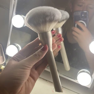 Jumbo Powder Brush - Säljer en supermjuk och fluffig sminkborste, perfekt för att applicera puder jämnt över ansiktet. Den har ett stilrent beige handtag och täta, vita borst som ger en lyxig känsla. Borsten är märkt med '101 Jumbo Powder Brush' och är idealisk för både vardags- och festsminkningar märket är FLEAR , nypris 220kr.