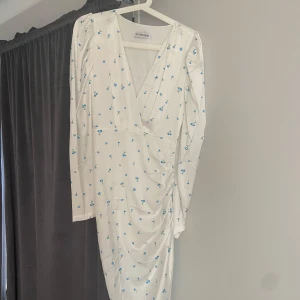 Vit blommig klänning från ADOORE - Säljer en superfin vit klänning från ADOORE med blå blommor. Den har en v-ringad design och långa ärmar. Klänningen är i ett lätt och bekvämt material, perfekt för vår och sommar. Observera att det finns en liten skada på ena sidan, se bild för detaljer.