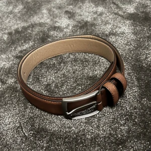 Brunt skärp i skinn - Säljer ett snyggt brunt skärp i skinn och mocka med en stilren silverfärgad spänne. Perfekt för att ge en klassisk touch till vilken outfit som helst. Skärpet har flera hål för justerbar passform och är i nyskick. Passar både till vardag och fest!