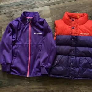 Lila och röd dunväst från Everest - Säljer en cool lila och röd dunväst från Everest. Västen har en snygg färgkombination med röd upptill och lila nedtill. Den är perfekt för höst och vinter och har knappar framtill. Västen är i bra skick och redo för nya äventyr!