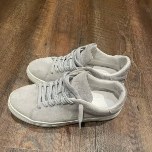 Grå sneakers i mocka - Säljer ett par stilrena grå sneakers i mocka. De har en klassisk design med snörning och en bekväm passform. Perfekta för både vardag och festliga tillfällen. Skorna är i nyskick och redo att användas