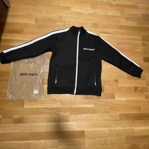 Svart jacka från Palm Angels - Säljer en snygg svart jacka från Palm Angels. Den har vita ränder längs ärmarna och en dragkedja framtill. Perfekt för en casual look eller när du vill ha något stilrent och bekvämt. Jackan är i nyskick och kommer med originalförpackning.