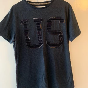 Mörkblå t-shirt med US-tryck - Säljer en snygg mörkblå t-shirt med stort 'US' tryck på framsidan. T-shirten är i bomull och har en normal passform. Perfekt för vardagsbruk eller en avslappnad dag ute. Den är kortärmad och i bra skick!