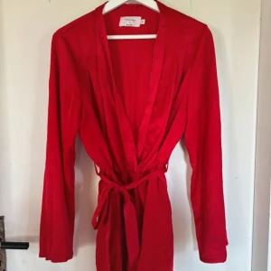 Röd playsuit från NA-KD - Säljer en snygg röd playsuit från Hanna Licious x NA-KD. Den har en djup v-ringning och är gjord i satinliknande polyester. Playsuiten har långa ärmar och ett knytband i midjan för en smickrande passform. Perfekt för festliga tillfällen!