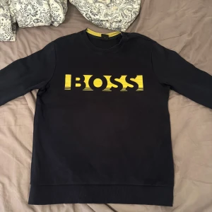 Svart tröja från Boss - Säljer en snygg svart tröja från Boss med ett gult tryck på framsidan. Tröjan har långa ärmar och en bekväm passform. Perfekt för både vardag och fest!