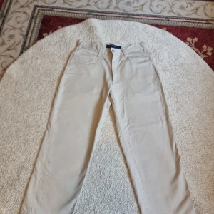 Beige byxor från Request - Säljer ett par snygga beige byxor från Request, exklusivt för Kappahl. De är tillverkade i 100% bomull vilket gör dem både bekväma och hållbara. Perfekta för en avslappnad men stilren look. Byxorna har en klassisk design med knappar och dragkedja.