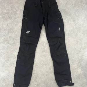 Svarta byxor från RevolutionRace - Säljer ett par svarta byxor från RevolutionRace i storlek M. Byxorna har flera praktiska dragkedjefickor och är utrustade med RECCO-reflektorer för extra säkerhet. De är perfekta för utomhusaktiviteter och har en bekväm passform med elastisk midja.