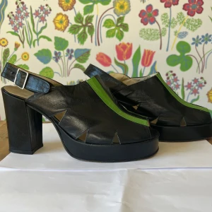 Svarta klackskor med platå - Snygga och unika klackskor från Arcadia i svart och grönt. Skorna har en öppen tå och en bred rem över foten med en justerbar spänne vid hälen. Perfekta för en utekväll eller fest!