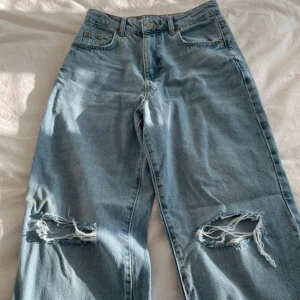 Ljusblå jeans med slitningar - Säljer ett par ljusblå jeans i 90-talsstil med straight passform. De har coola slitningar på knäna som ger en avslappnad look. Midjan är midwaist och de är i storlek 25/32. Perfekta för en casual outfit!