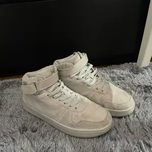 Säljer ett par snygga beige sneakers i mocka. De har en hög modell med snörning och kardborreband upptill för extra stöd. Skorna är i bra skick med lite slitage på sulorna. Perfekta för både vardag och festliga tillfällen!