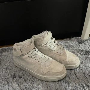 Beige sneakers i mocka - Säljer ett par snygga beige sneakers i mocka. De har en hög modell med snörning och kardborreband upptill för extra stöd. Skorna är i bra skick med lite slitage på sulorna. Perfekta för både vardag och festliga tillfällen!