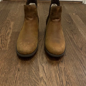 Bruna boots från UGG - Säljer ett par snygga bruna boots från UGG i skinn. De har en klassisk design med elastiska paneler på sidorna för enkel på- och avtagning. Perfekta för höst och vinter med en robust sula som ger bra grepp. Använda men i bra skick, redo för nya äventyr!