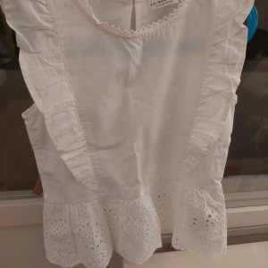 Vit blus från LC Waikiki - Söt vit blus från LC Waikiki för tjejer i storlek 8-9 år. Blusen har fina volangdetaljer och broderade spetsar vid ärmslut och nederkant. Perfekt för sommarens alla tillfällen!