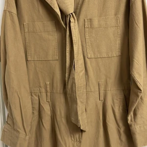 Brun khaki jumpsuit från H&M - Säljer en jumpsuit från H&M i storlek 34. Den har långa ärmar och ett knytbälte i midjan för en stilren look. Perfekt för höst och vår! Passar även storlek 38 