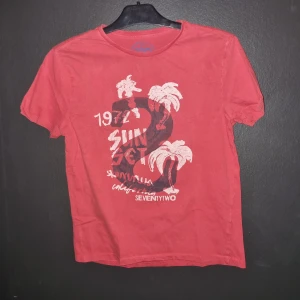 Röd t-shirt från 72D Jeanswear - Säljer en röd t-shirt från 72D Jeanswear i storlek medium. Den har ett coolt tryck med palmer och texten '1972 Sunset' på framsidan. 