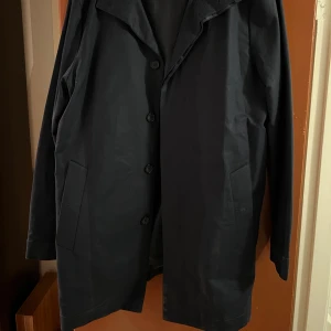 Svart kappa från Pier One - Säljer en stilren svart/mörkblå kappa från Pier One i storlek L. Den knappar framtill men syns inte väl stängd. Perfekt för höst och vår, med en klassisk design som passar till alla tillfällen. Kappan är i mycket bra skick. 