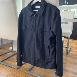 Svart jacka från These Glory Days - Säljer en stilren svart jacka från These Glory Days i storlek medium. Jackan har en klassisk krage och dragkedja framtill. Perfekt för höst och vår med sin enkla och tidlösa design. Den är i mycket bra skick och passar till de flesta outfits. 