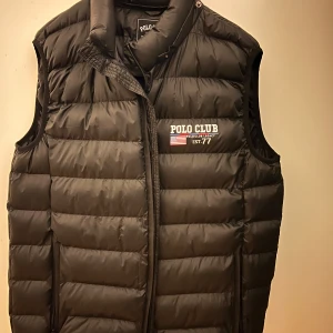 Svart dunväst från Polo Club - Säljer en svart dunväst från Polo Club i storlek XL. Västen har en quiltad design med dragkedja och en liten logga på bröstet. Perfekt för höst och vinter för att hålla värmen utan att kompromissa med stilen. Passar bra till både vardag och fest.