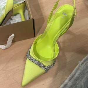 Neongula klackskor med strassdetaljer - Snygga neongula klackskor från Zara i nytt skick. De har en spetsig tå och en elegant klack. Skorna är dekorerade med glittrande strassdetaljer som ger en extra touch av glamour. Perfekta för festliga tillfällen!