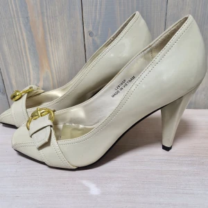 Beige pumps med gulddetalj - Snygga beige pumps i skinn med en elegant gulddetalj framtill. De har en spetsig tå och en stabil klack som ger en klassisk look. Perfekta för både fest och mer formella tillfällen. Skorna är i fint skick och redo att användas för att lyfta din outfit.