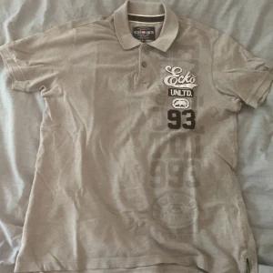 pikétröja från Ecko Unltd - Säljer en snygg grå pikétröja från Ecko Unltd i storlek L. Tröjan är kortärmad och har en klassisk krage med knappar. Den är gjord av 100% bomull och har coola tryckdetaljer med texten 'Ecko UNLTD' och siffran '93' på framsidan. Skriv om ni vill ha fler bilder eller om ni har frågor!! Priset är diskuterbart!