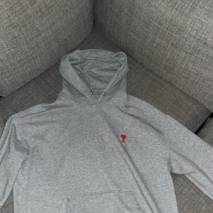 Grå hoodie med röd detalj - Säljer en stilren grå hoodie i bomull med en liten röd broderad detalj på bröstet. Perfekt för höst och vinter med sin mysiga känsla. Den har en klassisk passform och en stor ficka framtill. Perfekt för både vardag och chill! 🧢