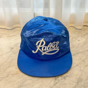 Blå keps med broderad text - Säljer en cool blå keps med texten 'Rabbit' broderad framtill. Kepsen har en glansig finish och en justerbar rem baktill för perfekt passform. Perfekt för en avslappnad stil eller som statement piece. Lite använd men i bra skick!    🧢 Jag har flera kepsar till salu på min profil och lägger upp nya modeller regelbundet. 