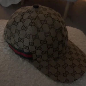 Beige Guccikeps  - Snygg beige keps från Gucci med det klassiska gg mönstret. Kepsen har en justerbar rem baktill och en röd och grön randdetalj på sidan. Perfekt för att ge din outfit en lyxig touch. Kepsen är aldrig använd, pris kan diskuteras vid snabbaffär 