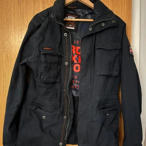 Svart jacka från Superdry - Säljer min killes jacka från Superdry. Jackan har flera fickor och en dragkedja framtill. Perfekt för både höst och vår. Nypris är mellan 2500-3000 och jackan är i nyskick. 