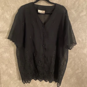 Svart blus från Dorothy Perkins - Elegant svart blus från Dorothy Perkins med v-ringning och knappar framtill. Blusen har kortärmade ärmar och vackra broderade detaljer längs kanterna. Perfekt för en stilren look.