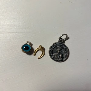 Tre berlocker i olika stilar - Säljer tre unika berlocker: en silverfärgad medaljong med religiöst motiv, en guldhästsko och en blått öga med guldkant. Perfekta för att skapa ett personligt halsband eller armband.