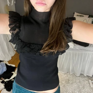 Svart ärmlös topp med volangdetaljer - Snygg svart ärmlös topp med volangdetaljer runt ärmhålen och halsen. Perfekt för en stilren look.