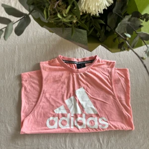 Rosa ärmlös topp från Adidas - Säljer en snygg rosa ärmlös topp från Adidas med deras ikoniska logga i vitt framtill. Perfekt för träning eller en avslappnad dag. Toppen är gjord i mjukt bomullsmaterial.