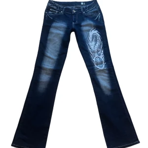 y2k vintage jeans med drakmotiv crazy age  - Snygga mörkblå jeans från Crazy Jeans med ett coolt drakmotiv på framsidan. Jeansen har en bootcut-stil och är dekorerade med broderade detaljer på bakfickorna. Strl L på lapp men mer en S i storlek. Midjemått: 36 cm rakt över, Innerbenslängd 84 cm. Är stretchiga. 💞