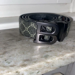 Svart Burberry Bälte - Snyggt svart bälte med ett diskret mönster och ett stilrent metallspänne. Perfekt för att ge din outfit en extra touch. 